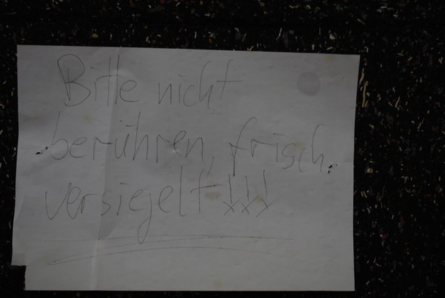 In diesem Tor vor einem Geräteraum klaffte bis vor Kurzem ein Loch, das nun mit einer Platte versehen wurde. Darauf steht in Krakelschrift der wohl ironisch gemeinte Satz: „Bitte nicht berühren, frisch versiegelt!!!“