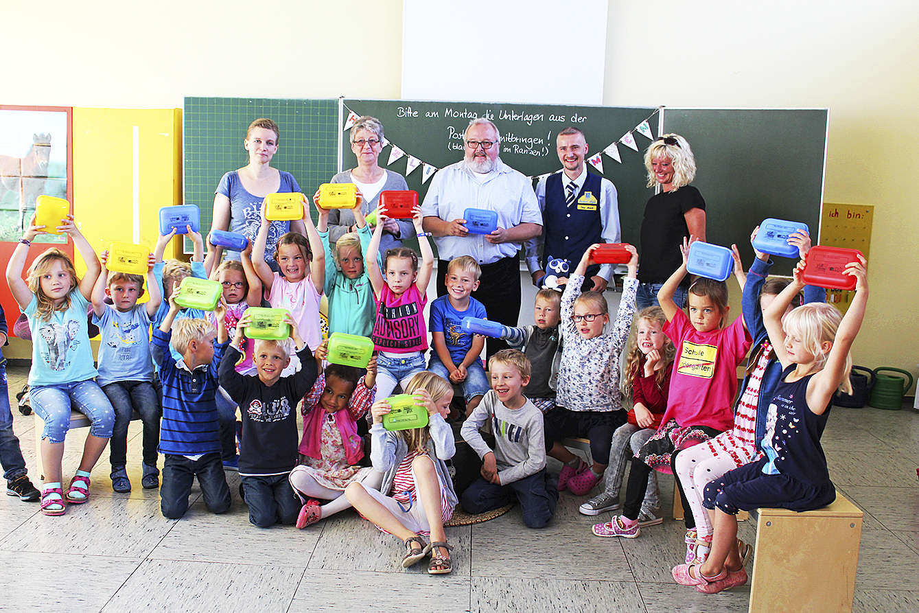Lehrerin Jessica Fürch (hinten, v. li.), Giesela und Rolf Wiese, Timo Jänsch (Edeka Meyer) und
Direktorin Ranga Hoffmann mit den Erstklässlern, die stolz ihre neuen Brotdosen zeigen