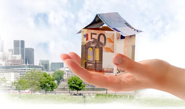 Ein Haus aus Eurobanknoten auf einer Hand. Im Hintergrund Gebäude  | Foto: adobestock@tesgro tessieri