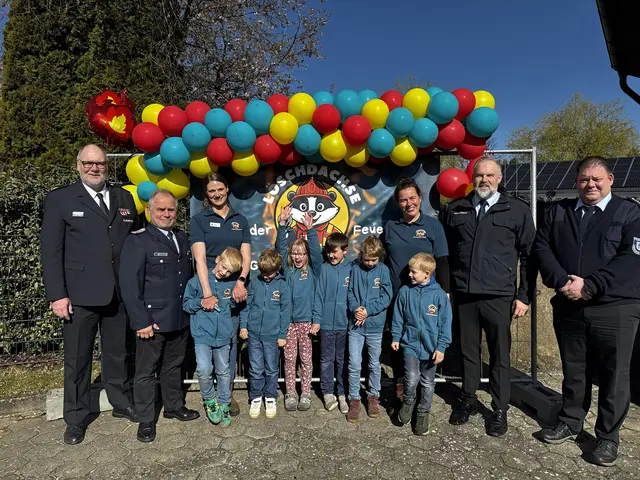 Sechs Erwachsene und sechs Kinder stehen vor bunten Luftballons und dem Logo der Kinderfeuerwehr