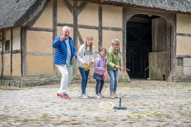 Älteres Ehepaar mit zwei Enkelinnen spielen Ringe werfen vor einem historischen Bauerngebäude.